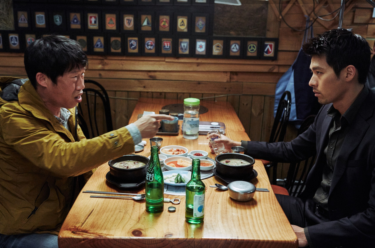 식당에서 유해진이 현빈에게 손가락을 가리키며 말하는 장면, 테이블에 소주병과 국밥이 놓여 있다