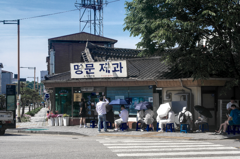 흰색 바탕에 파란 글씨로 된 명문제과 간판. 그 앞에 우산으로 햇빛을 가리고 대기 중인 고객들