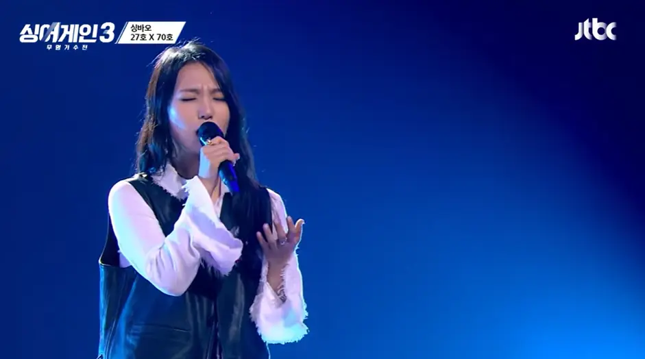 싱어게인3- 흰셔츠 검정색 조끼를 입고 웨이브진 긴머리로 오른손에 마이크를 잡고 서서노래하는 27호 상반신