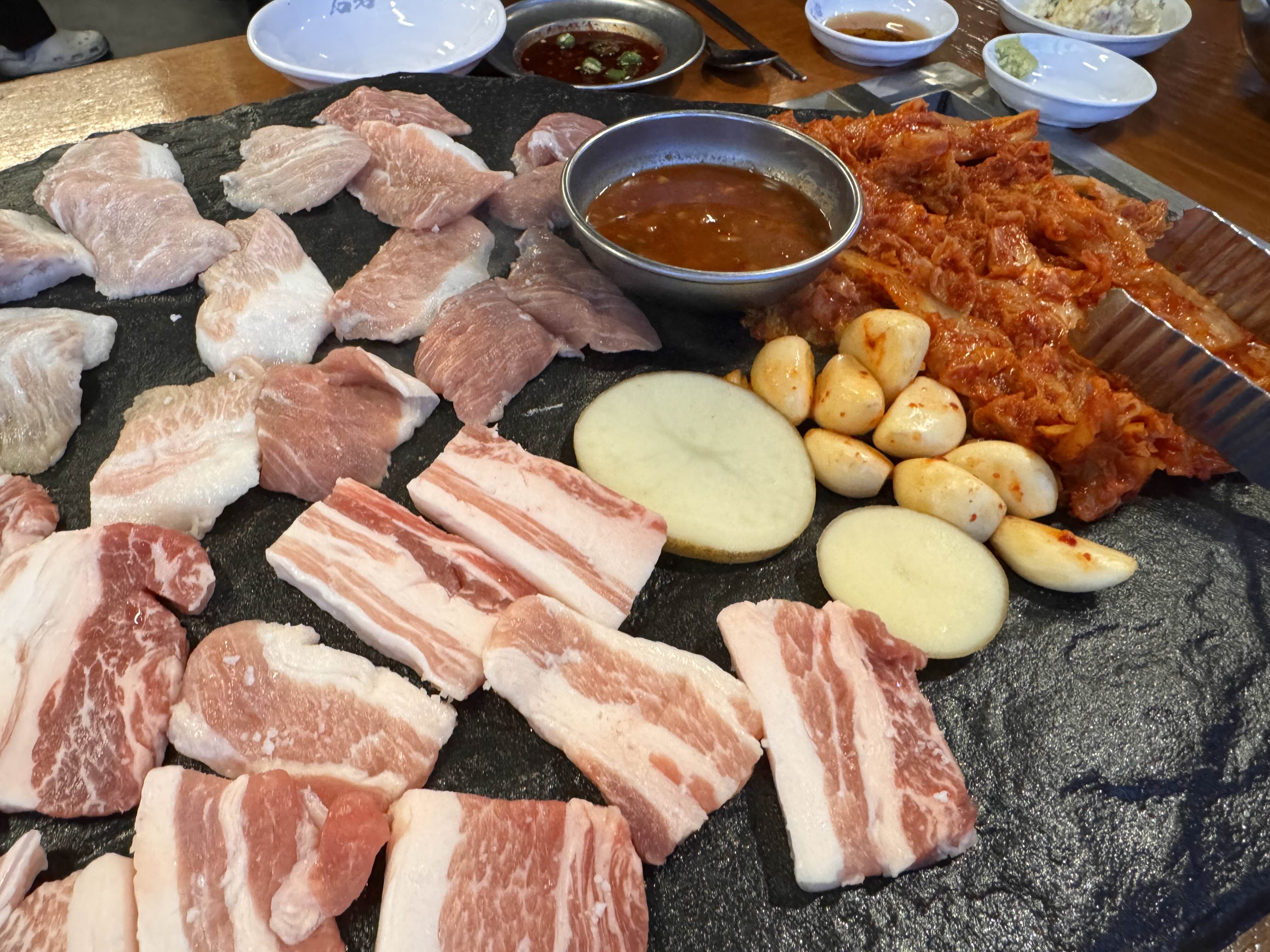 돌판 위에 가지런히 놓인 선홍빛 생삼겹살과 항정살, 그리고 함께 구워 먹는 김치, 마늘, 감자가 어우러진 먹음직스러운 모습