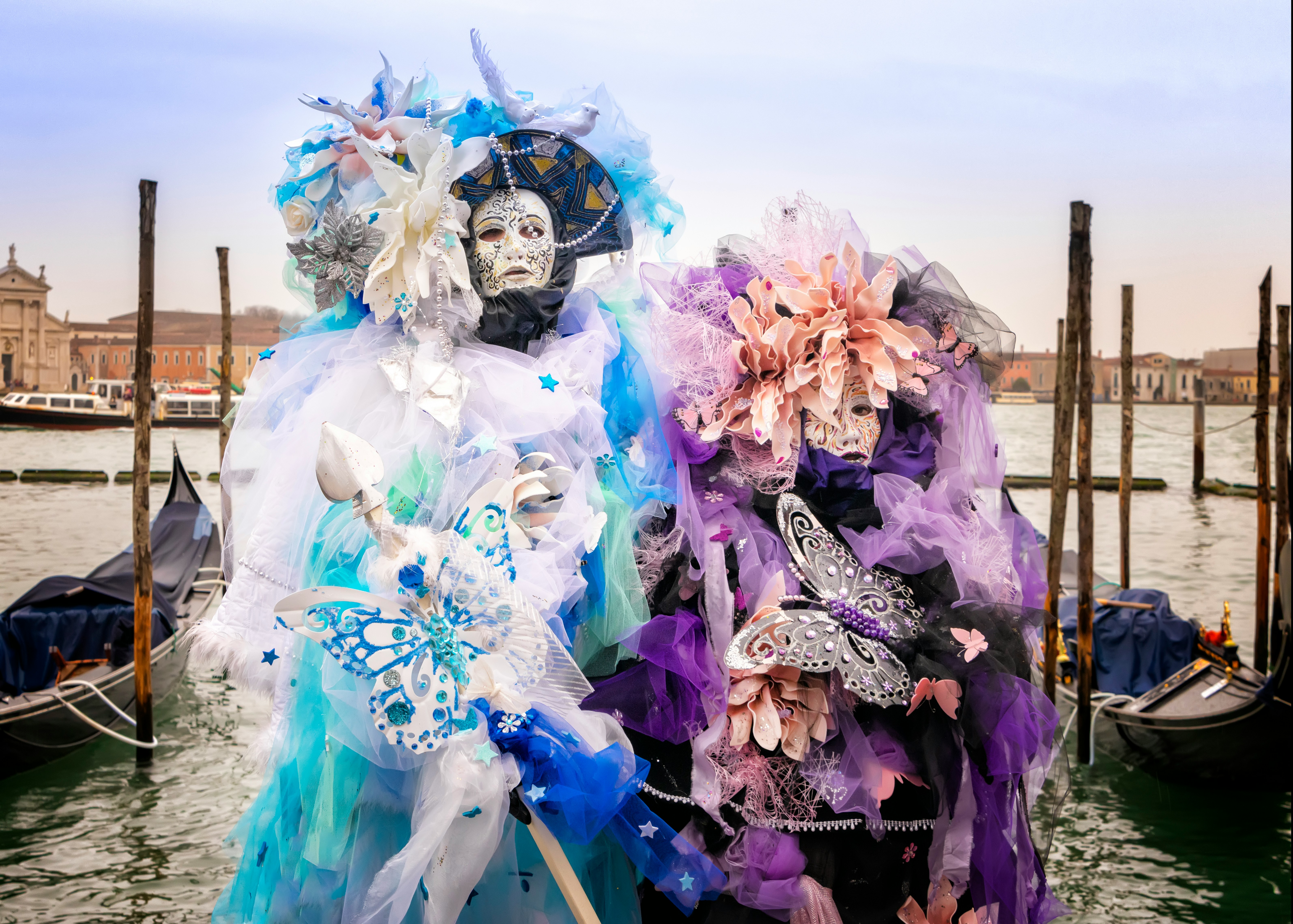 베네치아 카니발에서 화려한 가면과 의상을 입은 두 사람이 운하를 배경으로 서 있는 모습
