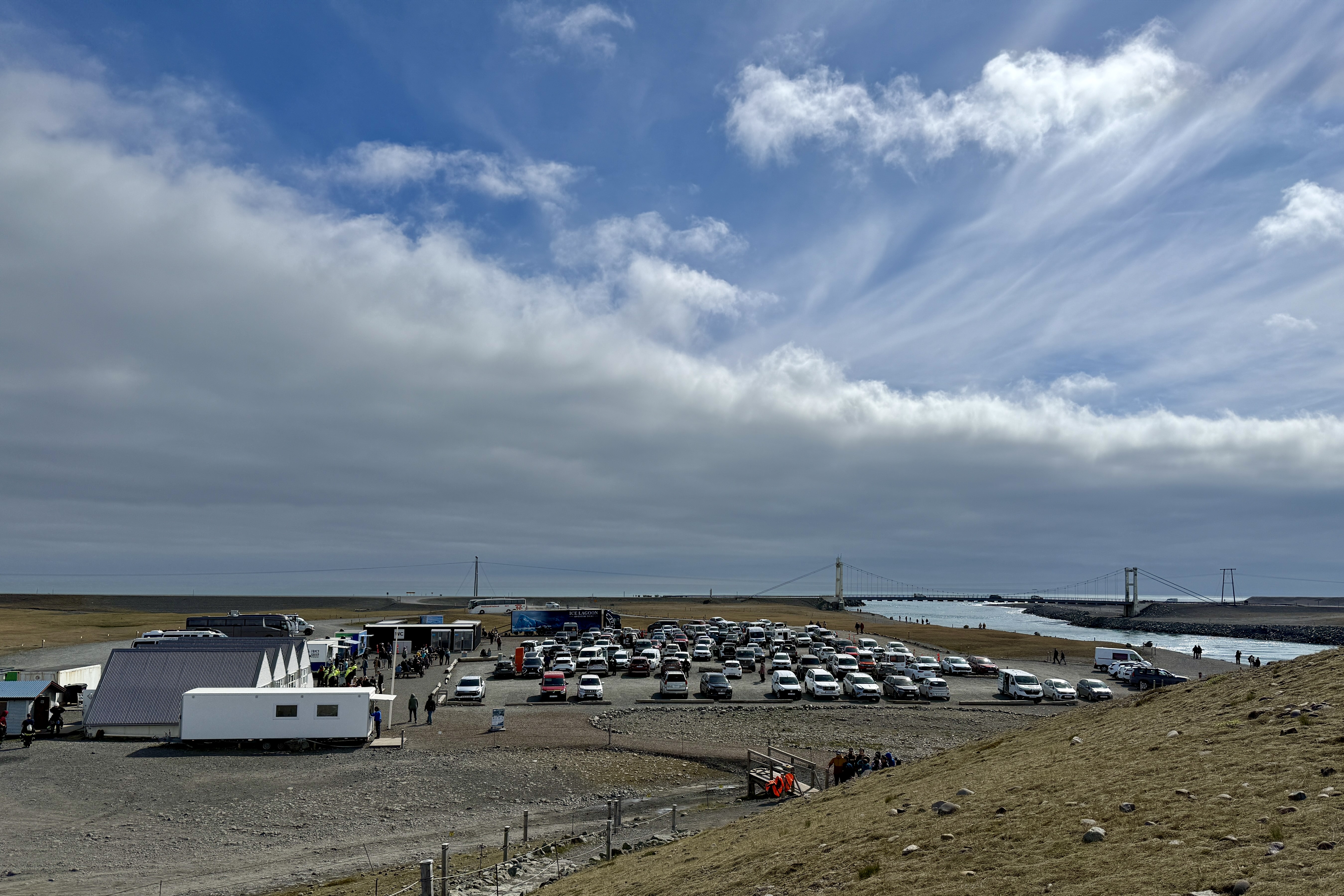 요쿨살론 주차장 전경. 많은 관광객 차량들이 가득 주차되어 있고, 흰색 건물들(푸드트럭과 편의시설)이 보임. 멀리 다리가 있고 주변은 황량한 자갈 지형으로 둘러싸여 있음. 아이슬란드 특유의 넓은 하늘과 구름이 인상적임.