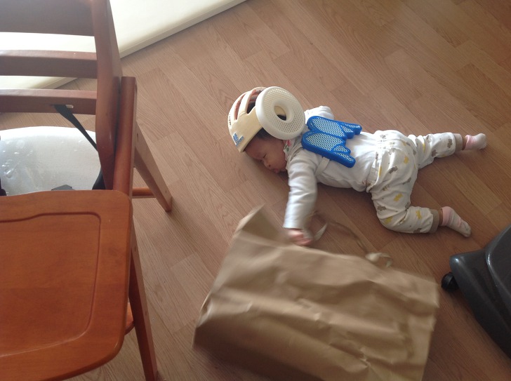 A baby wearing a BaileyBao safety helmet crawls under a chair toward an air purifier on a wooden floor, exploring the room safely.