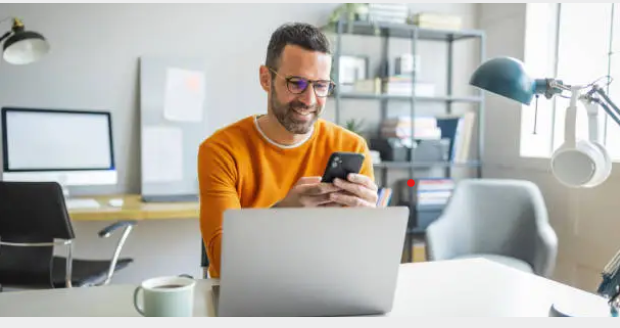 현대적인 사무실에서 노트북 앞에 앉아 스마트폰을 사용하며 미소 짓는 남성