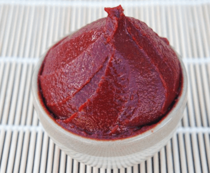 갈색 도기 그릇에 빨간 고추장이 한가득 담겨있고 그릇 바닥에는 갈색 나무 깔판이 있다.