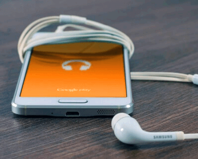 갈색 나무 책상 위에 주황색 바탕화면을 가진 휴대폰이 있고 하얀색 이어폰이 휴대폰을 둘둘 말고 있다.