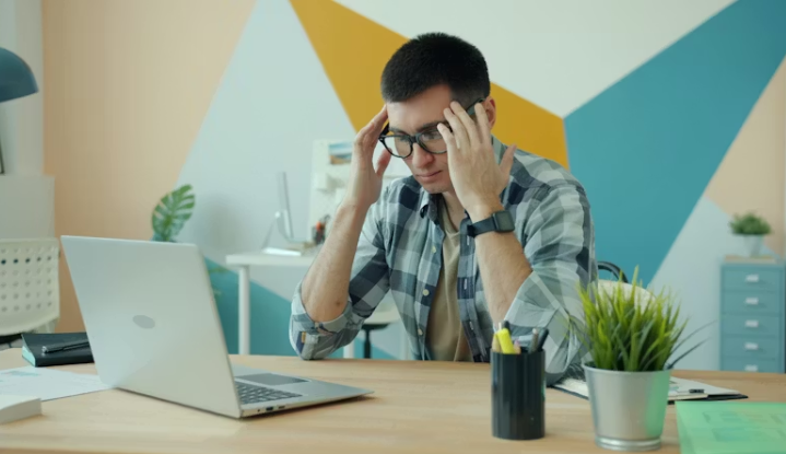 책상 앞에서 머리를 짚고 앉아 있으며 옆에 커피잔이 놓인 채 피로를 느끼는 모습