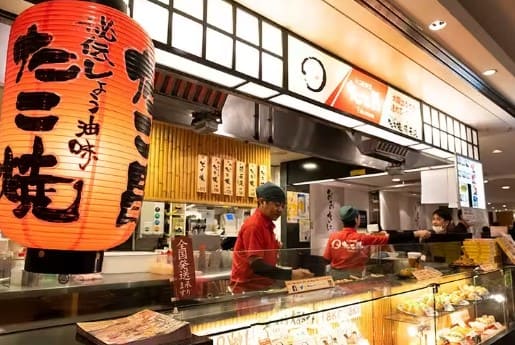 일본식 등불에 가게 이름이 적혀 있고 그 뒤로 가게 외관과 빨간색 옷을 입을 사람둘이 서 있다.