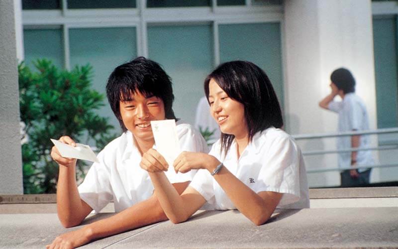 영화 '세상의 중심에서 사랑을 외치다'에서 교복을 입은 소년과 소녀가 사진을 보며 웃고 있는 장면