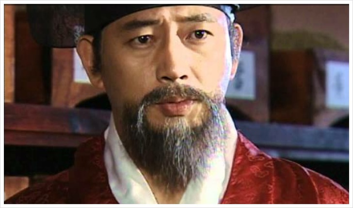 드라마 허준 장면: 붉은 관복을 입은 허준이 내의원 의관으로 앉아 진지한 표정으로 환자를 바라보는 모습