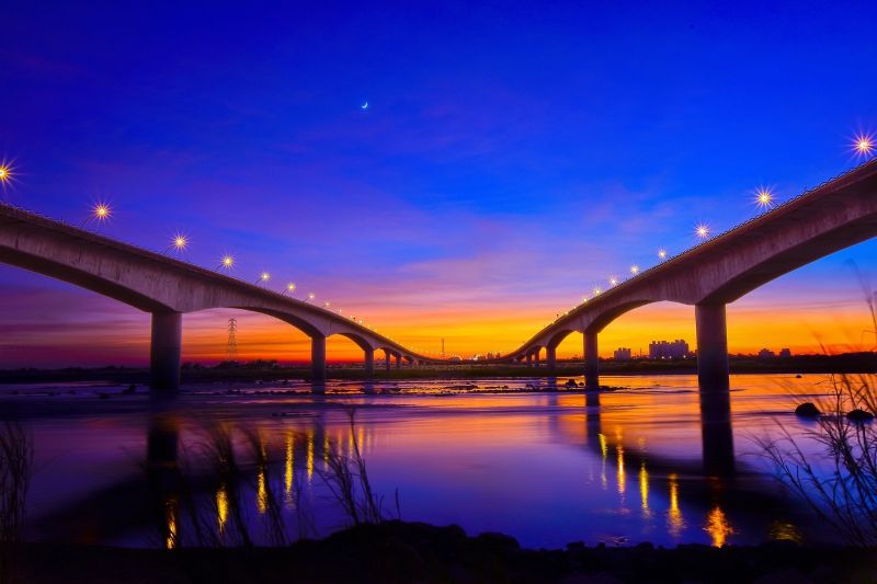 “대만의 강 위를 가로지르는 다리에 불이 밝혀지고, 붉게 물든 저녁노을과 파란 하늘이 어우러진 야경 전경”