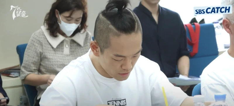 사람들이 앉아 있는 어느 회의실을 배경으로 짧은 사기컷 머리와 흰 반팔티를 입고 앉아 있는 드라마 7인의 탈출의 백익호 캐릭터