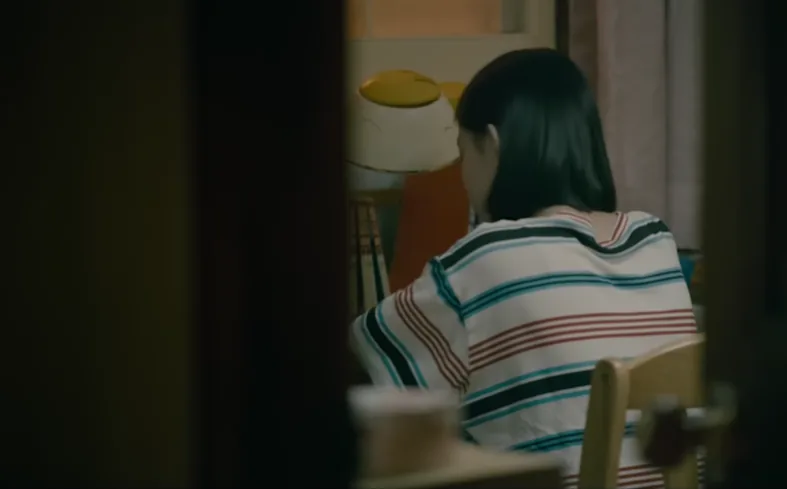 줄무늬 옷을 입은 소녀가 방 안 책상에 앉아 있는 영화 속 장면