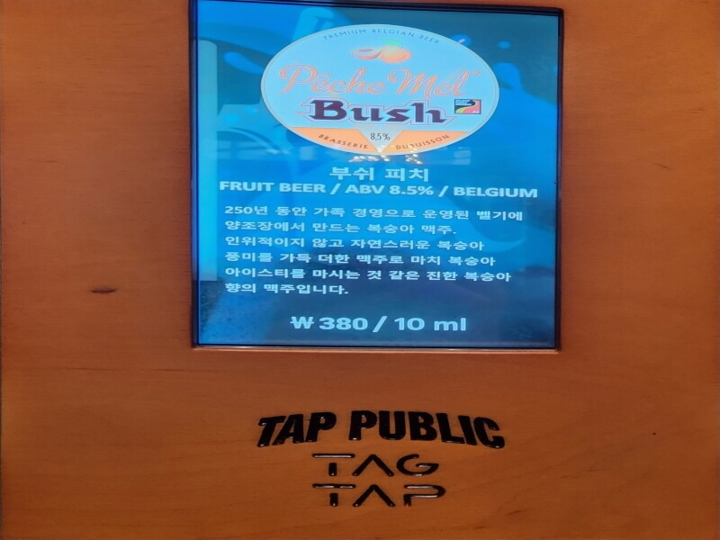 &#39;부쉬 피치&#39; 맥주에 대한 설명이 스크린에 떠 있음. &#39;250년 동안 가족 경영으로 운영된 벨기에 양조장에서 만드는 복숭아 맥주. 인위적이지 않고 자연스러운 복숭아 풍미를 가득 더한 맥주로 마치 복숭아 아이스티를 마시는 것 같은 진한 복숭아 향의 맥주&#39; 라는 설명이 쓰여 있음. 10ml에 380원.