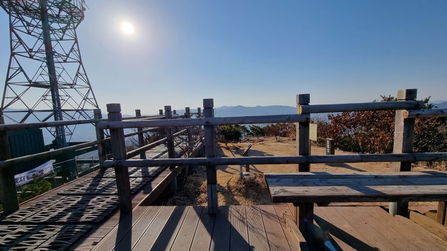 정상 전망대 난간 너머에 작고 검은 돌의 정싱석, 앞에는 나무판 장의자, 왼쪽은 내려가는 데크길, 타이어 고무 안전판이 깔려 있음, 왼쪽은 송신탑, 역광, 푸른하늘,