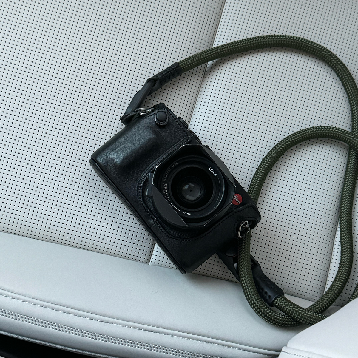 자동차 시트 위에 놓인 검은색 라이카 Q. 레늦 후드 끝부분에 미세한 흠집이 보이며, 상단에 빨간색 라이카 로고가 선명하게 드러난 정면 사진.