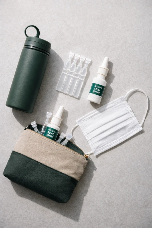 기내 건조함에 대비하기 위한 준비물인 재사용 텀블러, 일회용 무방부제 인공눈물 앰플, 등장성 비강 스프레이, 면 마스크가 작은 파우치와 함께 밝은 회색 배경 위에 정리되어 놓인 플랫레이 사진.