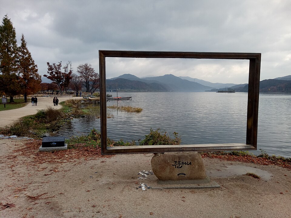 A large rectangular photo frame stands beside Dumulmeori&rsquo;s riverside path, framing calm water and distant hills under a cloudy sky.
