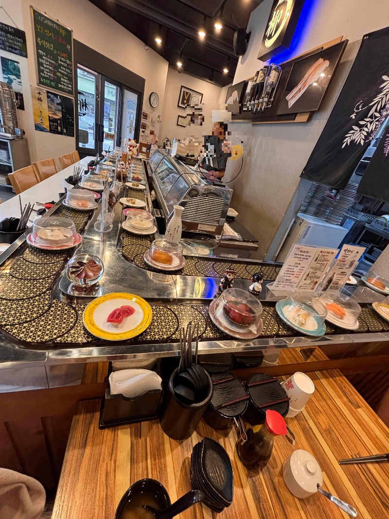 ํ์ ํ
์ด๋ธ๊ณผ ์
ฐํ๊ฐ ์๋ K-์ค์ ๋ ์คํ ๋ ๋ด๋ถ
Interior of a K-sushi restaurant with rotating plates and chef at the counter