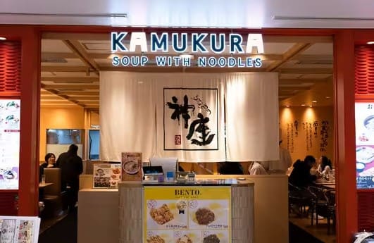 '카무쿠라' 라고 영어로 적혀 있는 가게의 입구 사진 뒤로 테이블에 사람들이 앉아 있다.