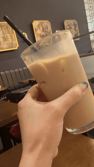 베질루트 시그니처 아이스 밀크티를 손으로 가볍게 흔드는 장면, 부드러운 색감의 음료와 카페 감성 인테리어 배경