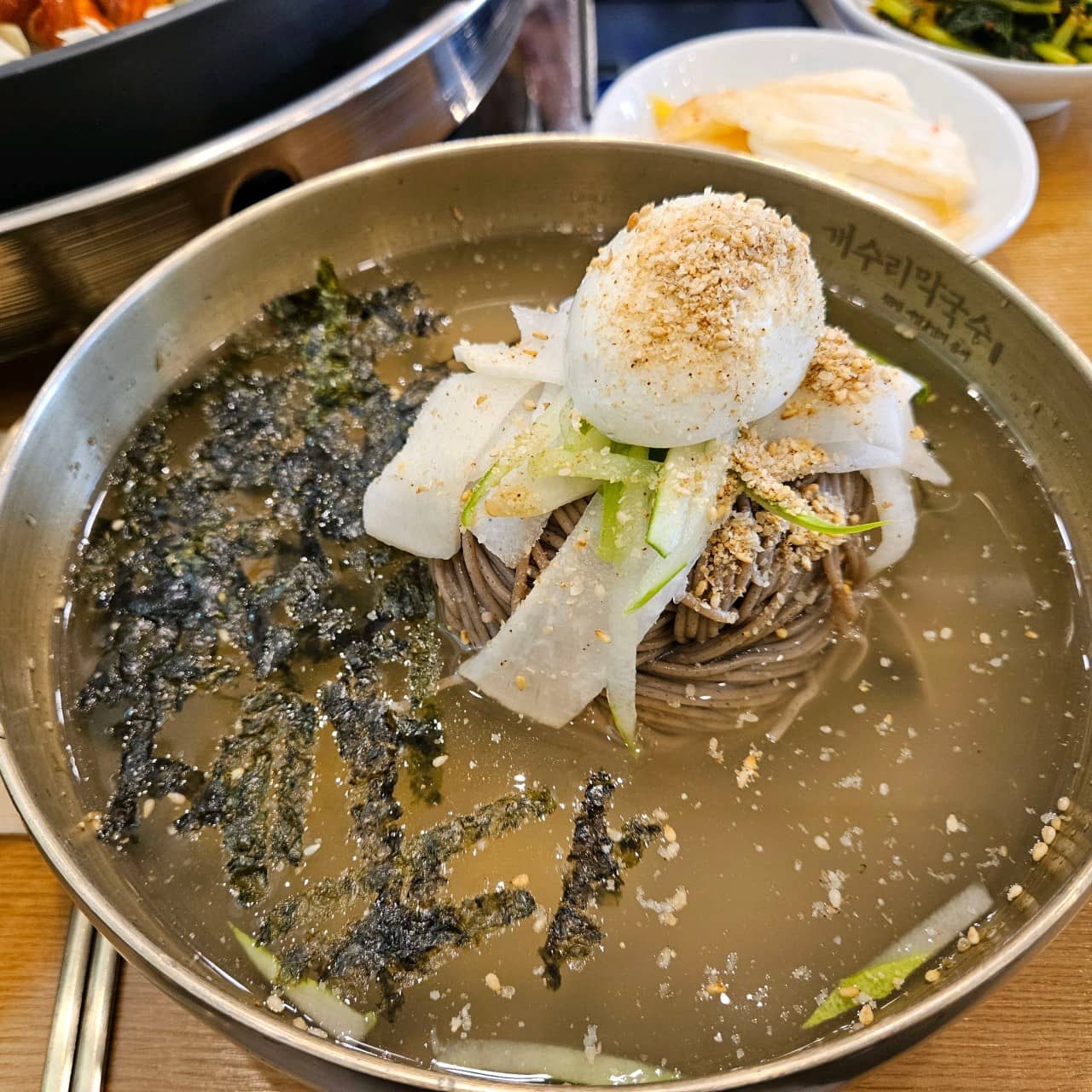 '개수리막국수' 물막국수 사진입니다. 냉면육수위에 김가루가 뿌려져 있고 메밀면 위에는 무절임, 오이, 삶은계란이 올려져있고 깨가 뿌려져 있습니다.