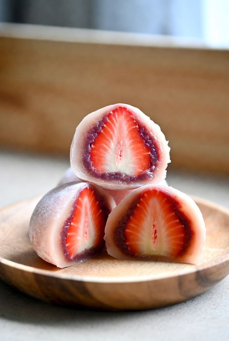 하얀 찹쌀떡 속에 선명한 붉은색의 싱싱한 딸기와 달콤한 팥소가 꽉 찬 단면을 보여주는 딸기 모찌. 미니멀한 나무 접시 위에 정갈하게 놓여 있으며, 부드러운 자연광이 내리쬐어 딸기의 과즙과 떡의 쫀득한 질감이 생생하게 느껴지는 전문적인 음식 사진