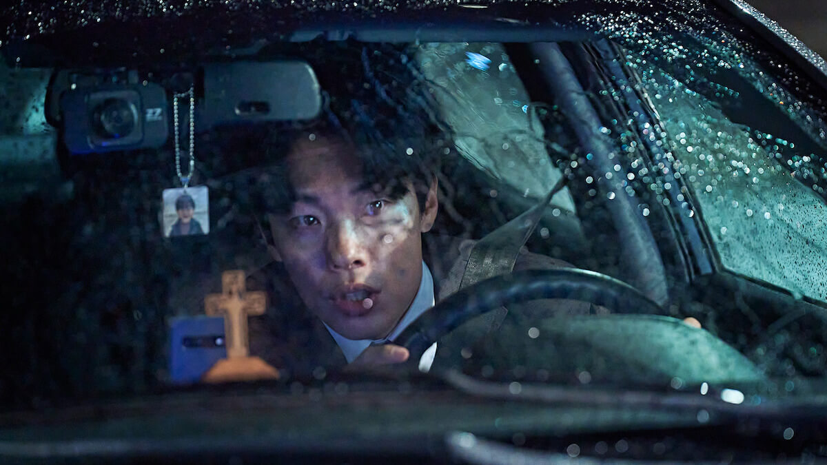 Revelations : 빗속 차량 안, 긴장한 주인공이 전방을 응시하고 있는 모습.
십자가 장식과 백미러 아래에 사진이 달린 목걸이가 있음.