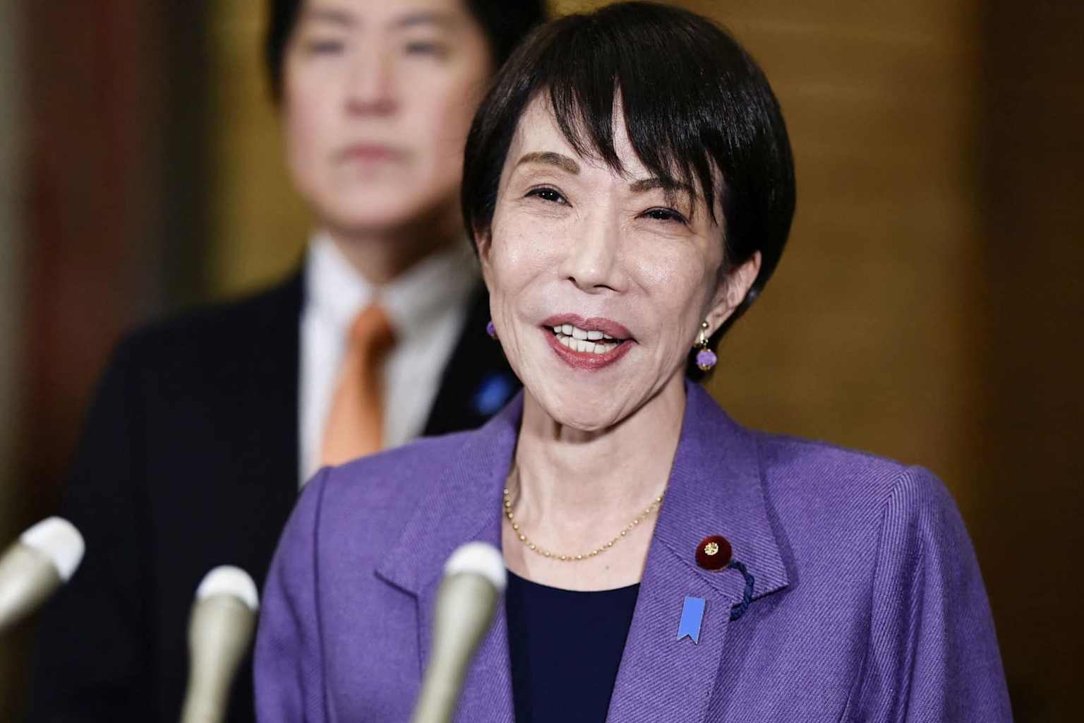 보라색 정장을 입은 다카이치 사나에 총리가 마이크 앞에서 말하고 있는 상반신 모습. 배경에는 정장을 입은 다른 정치인이 흐릿하게 보이며, 전형적인 일본 정치 발표 현장의 모습이다.