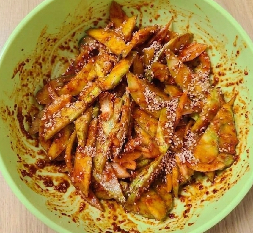 밝은 연두색 보울에 담긴 매콤한 빨간 양념의 오이무침과 그 위에 뿌려진 참깨.