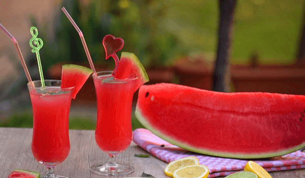 나무 식탁 위에 빨간 수박 조각이 있고 왼쪽에는 유리잔에 담긴 빨간 수박주스 두 잔이 있다. 유리잔에는 빨대가 꽂아있다.