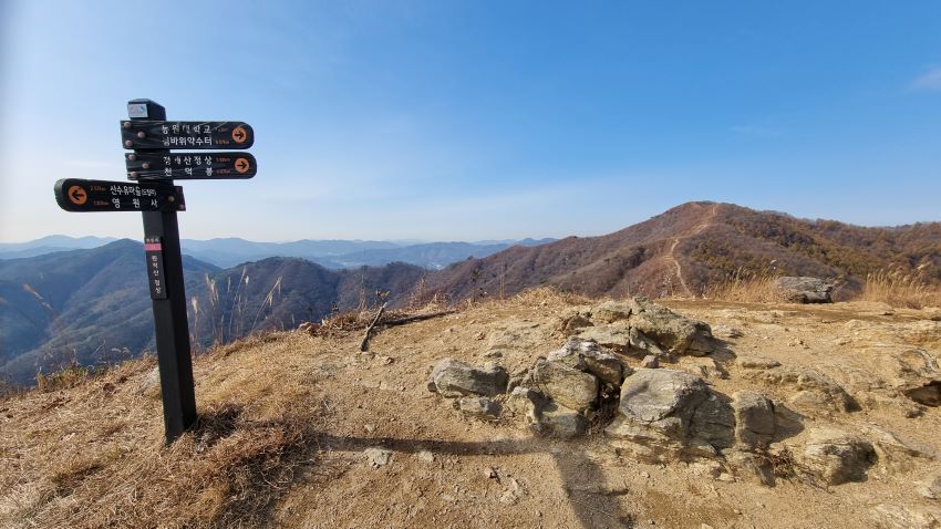 원적봉에서 바라본 천덕봉 가는 능선길, 왼쪽에 이정표, 길게 드리운 그람자, 푸른 하늘, 산 정상에 크지 않은 바위들,