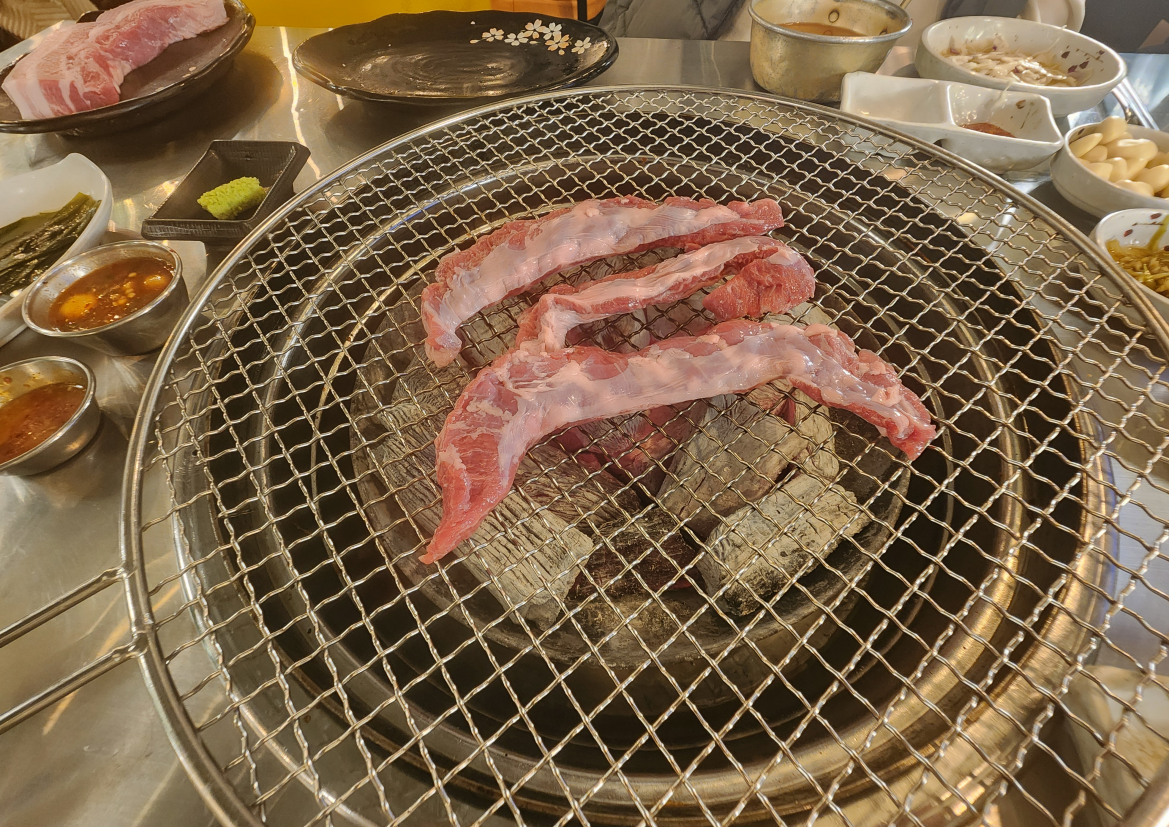 병점 민들레화로 숯불 화로 위에 생 돼지고기 부위가 철망 불판 위에 올려진 모습으로, 붉은 숯불 위에서 기름기 있는 고기가 아직 익기 전 상태로 펼쳐져 있고 주변에 양념장과 반찬 그릇이 함께 놓인 초기 굽기 단계 장면