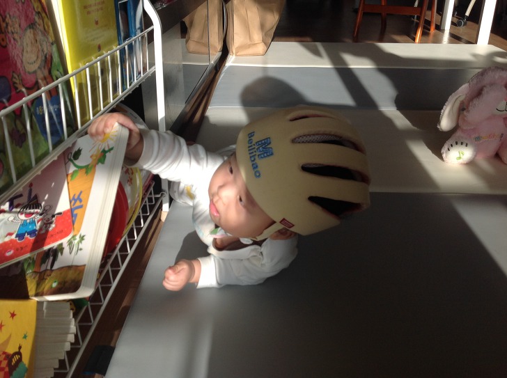 A baby wearing a cream-colored BaileyBao safety helmet reaches for a picture book on a low bookshelf.