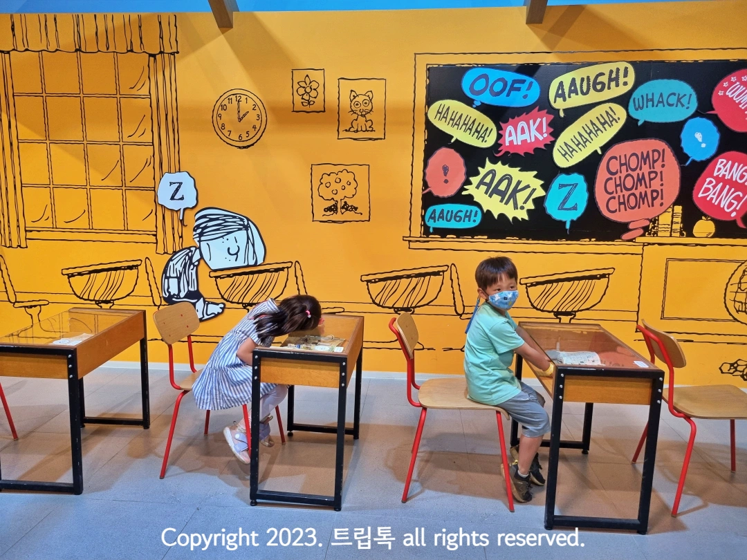 노란 벽에 학교 책상이 있고 아이2명이 앉아있다.