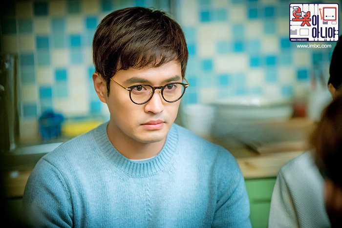 안경과 하늘색 스웨터를 착용한 홍백균이 정면을 바라보는 장면
