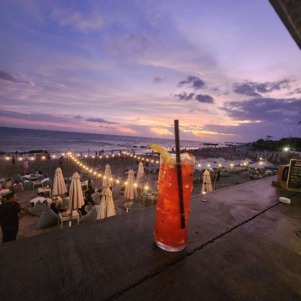 바다에는 노을, 바닷가에는 테이블과 조명, 선반에 칵테일 한 잔