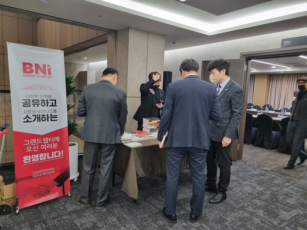 주간회의가 열리는 런던홀 로비 앞 테이블 주변에 여러 사람이 서 있고 비엔아이 그랜드챕터의 지식수준을 높여주는 책들이 테이블에서 대여를 기다리고 있습니다.