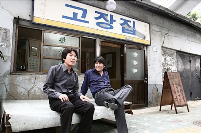 영화 《우아한 세계》에서 강인구와 친구가 허름한 고깃집 앞에서 담소를 나누는 장면. 조폭이라는 겉모습 뒤에 숨겨진 ‘평범한 가장’의 삶과 외로움이 조용히 스며든다.