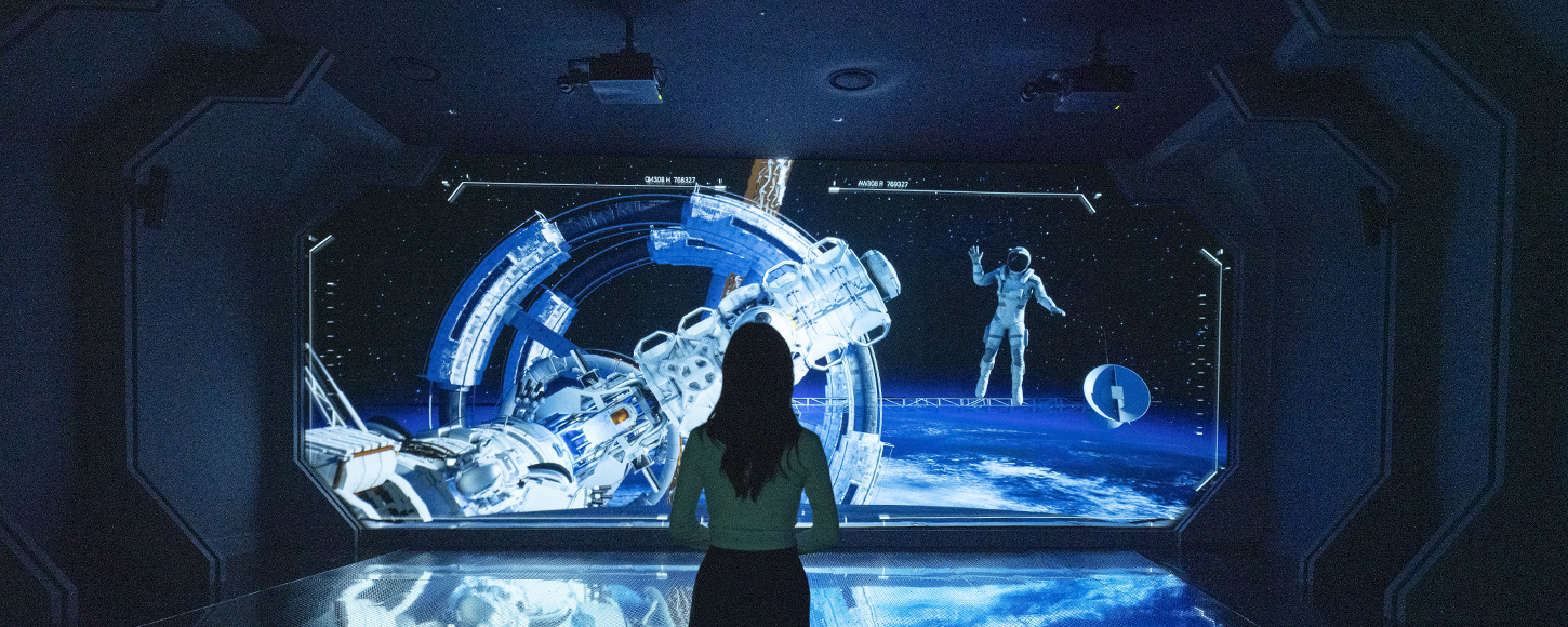 우주 공간을 형상화한 몰입형 전시관에서 한 관람객이 대형 스크린 앞에 서서 우주비행사와 우주정거장을 바라보고 있다.