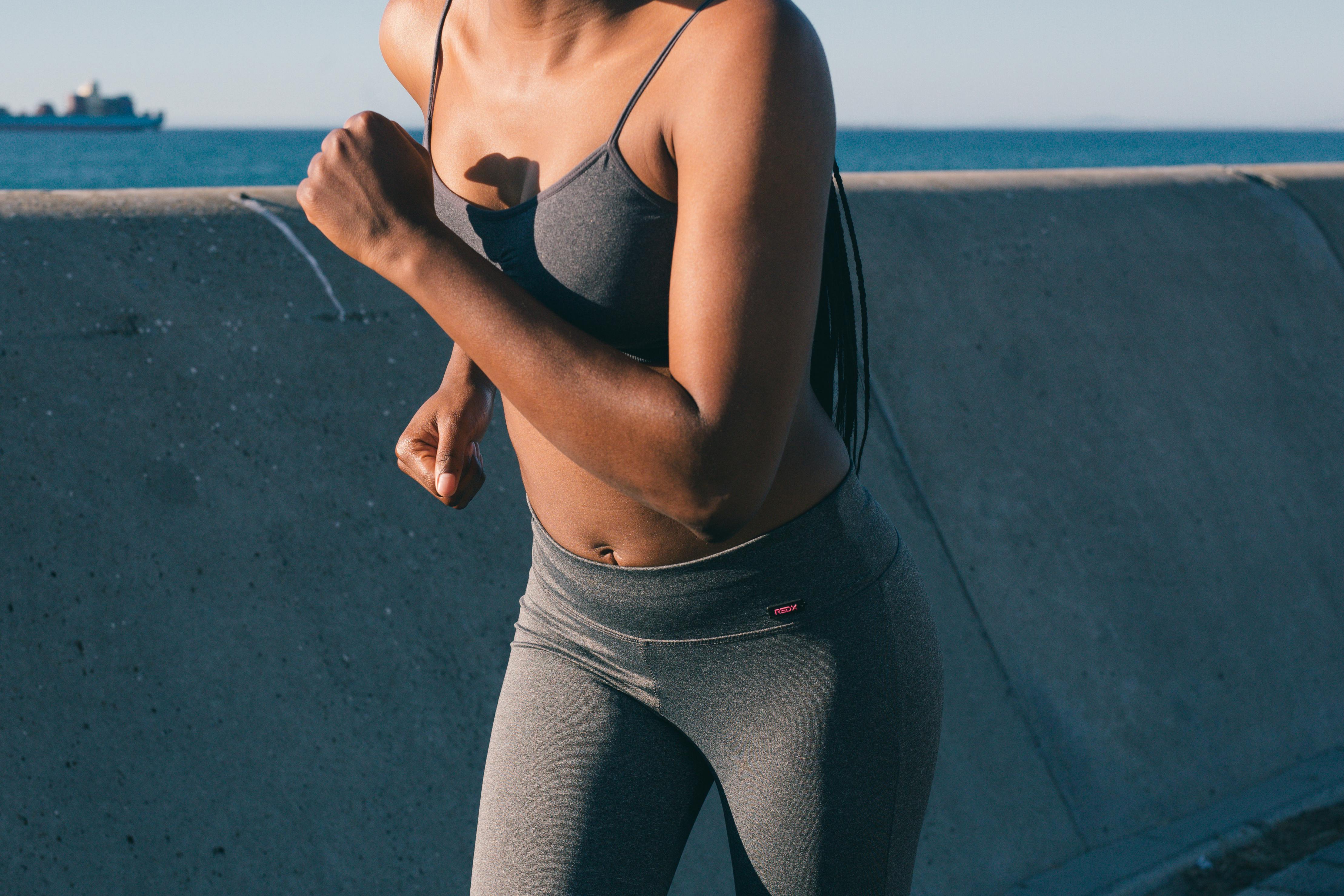 A mid-shot of a woman in gray athletic wear running along a concrete barrier with a body of water and a distant ship in the background