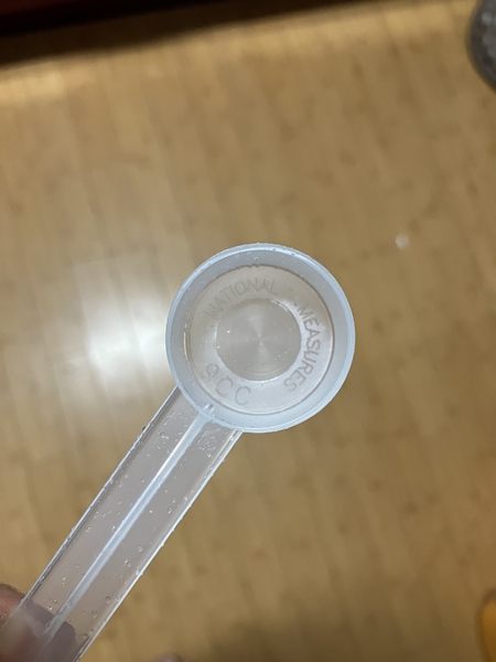 투명 플라스틱 재질의 긴 손잡이 9cc 계량 스푼, 손에 들고 바닥을 배경으로 촬영한 모습