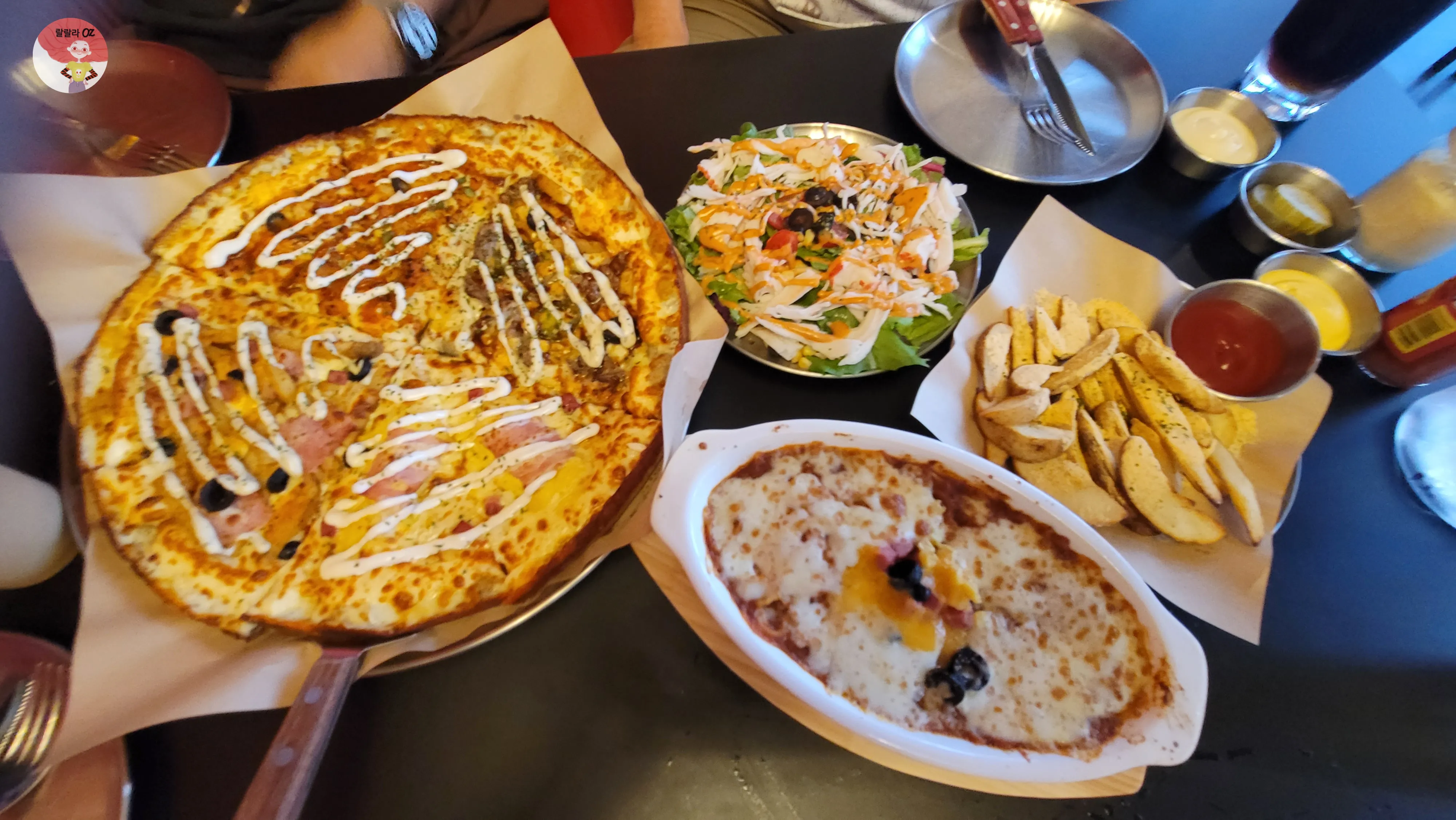 피자, 샐러드, 치즈오븐스파게티, 웨지감자, 음료가 함께 차려진 테이블에서 두 사람이 식사 중인 모습