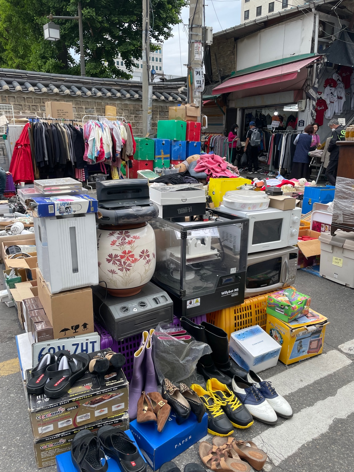 "Outdoor display at Dongmyo Market, showing secondhand home appliances, shoes, and clothing in plastic bins and cardboard boxes."