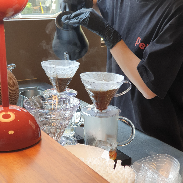 검은색 반팔티를 입은 바리스타가 검은색 장갑을 끼고 검은색 드립포트로 핸드 드립 중이다. 하리오 v60사용