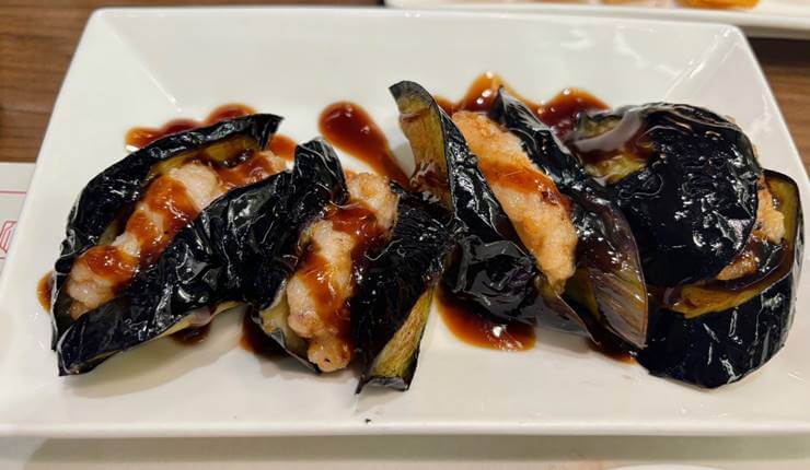 하얀 접시 위에 가지튀김 4개가 나란히 옆으로 놓여있다.
가지튀김은 가지 사이에 하얀 속이 있고 갈색 소스가 뿌려져 있다.