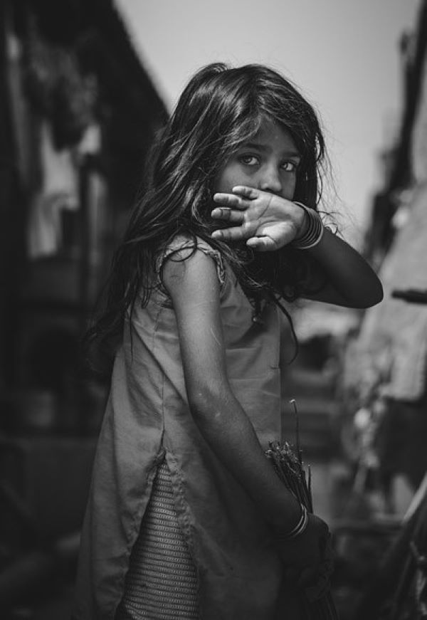 한 소녀가 어두운 골목에서 조용히 고개를 돌려 카메라를 바라보고 있는 흑백 이미지. 천진난만하면서도 슬픔이 깃든 눈빛이 어린아이의 보호받지 못한 현실을 상징함.