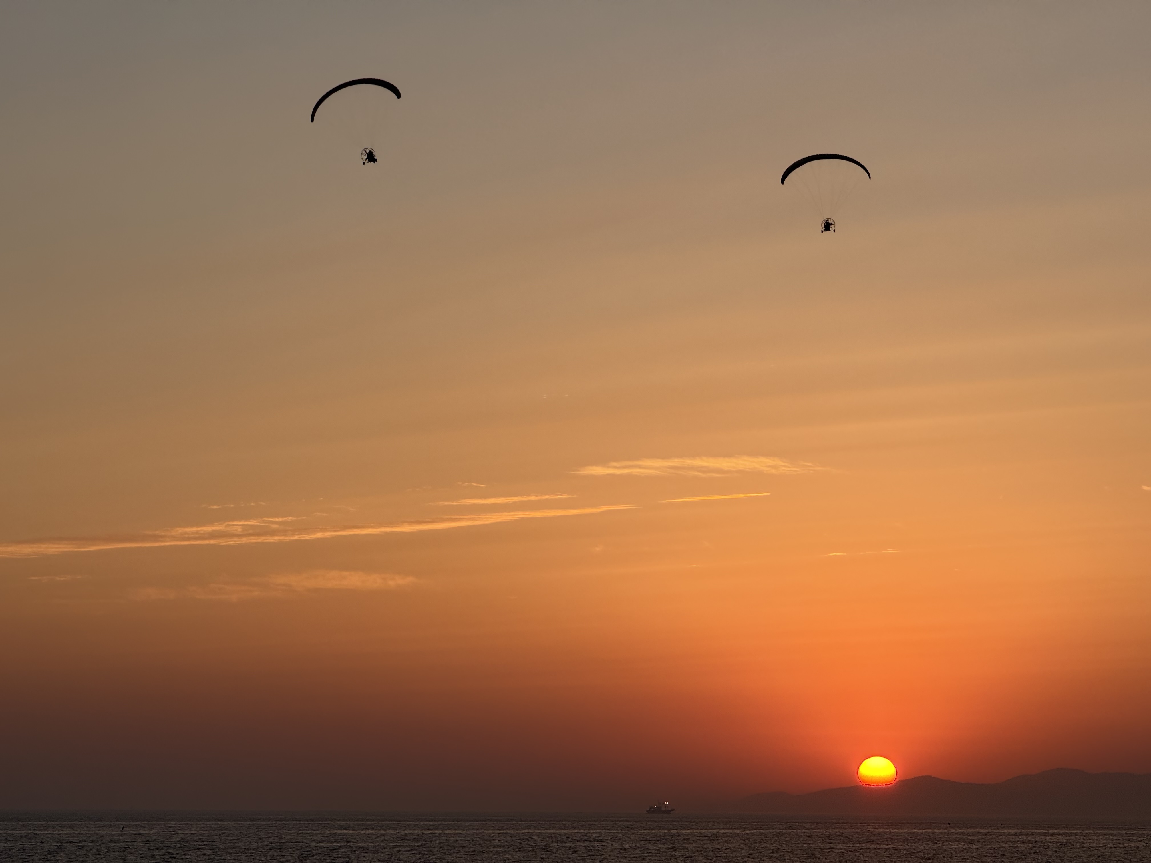 두 남자가 전동 패러글라이딩을 타고 해지는 바다 위를 날고 있습니다. 사진에서는 웃고 있는 눈동자와 눈썹 같습니다.