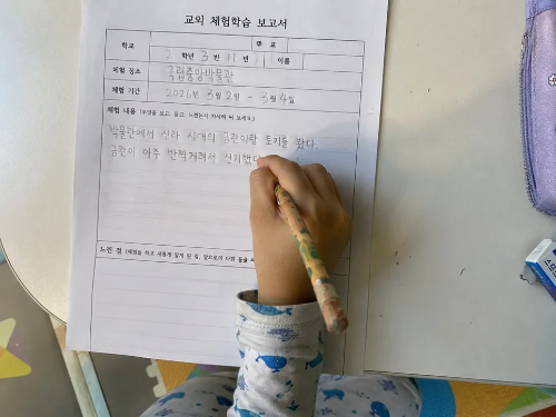 아이가 거실 테이블에서 연필을 쥐고 초등학교 교외체험학습 사후 보고서를 직접 손으로 작성하고 있는 모습