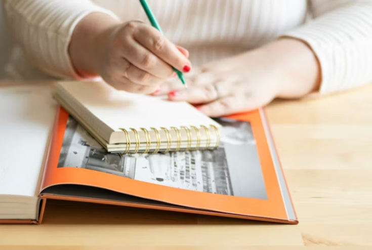 손으로 노트에 무언가를 적고 있는 모습. 책상 위에 펼쳐진 책과 노트가 함께 놓여 있다